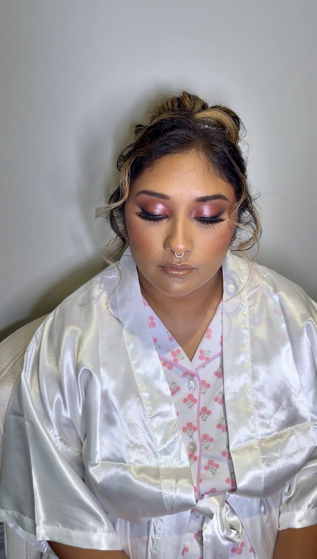 Bride getting ready in a satin robe with a pink shimmer eye, winged lashes, a glossy nude lip and hair in a curled updo