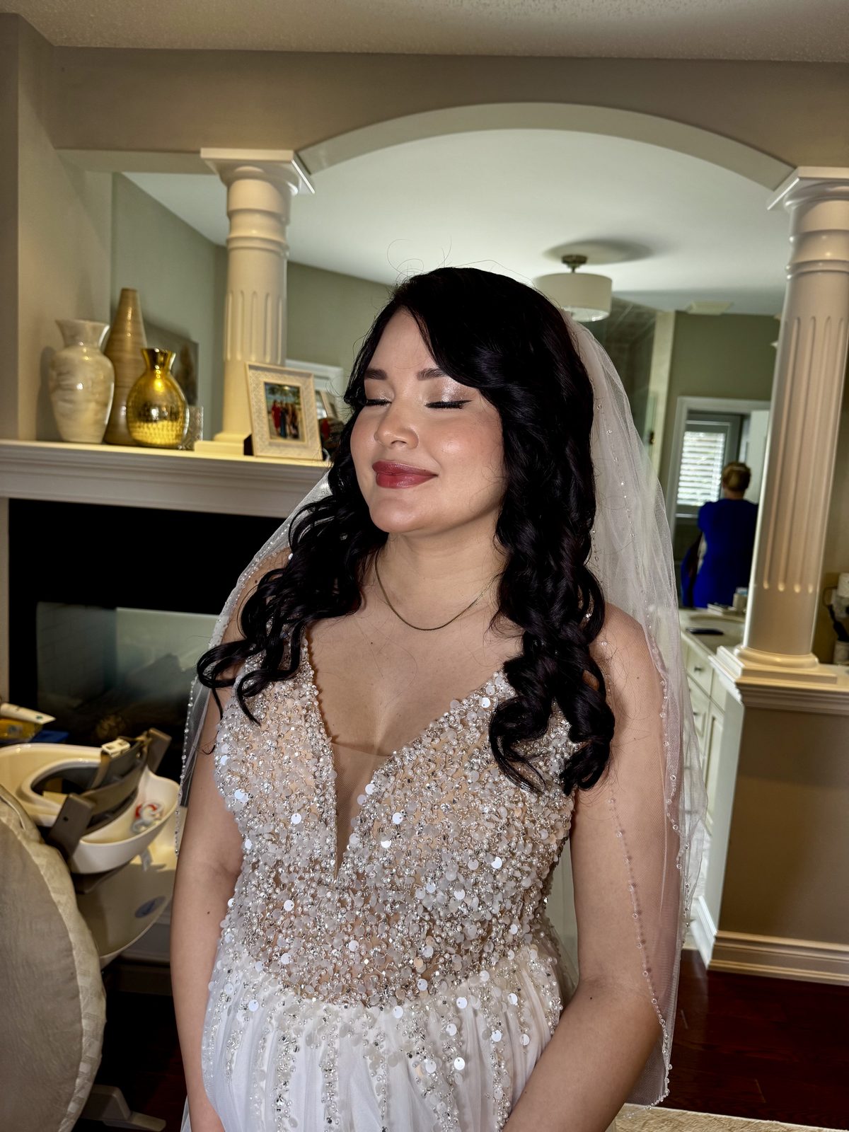 Bridal makeup and hair with glowing skin, soft neutral shimmer eyes and a rosy pink lip, worn with long dark curls and a beaded veil